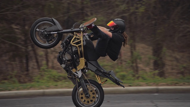 SLO MO，一名有着棕色长发绺和纹身的特技摩托车骑手，他的双腿伸直，同时跳起了一个前轮特技视频下载