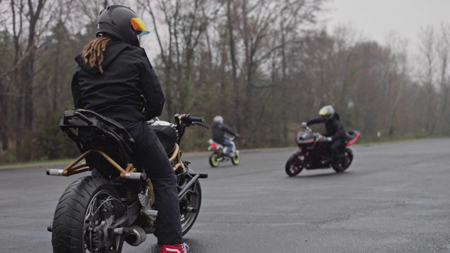一个留着棕色发辫的特技摩托车骑手坐在他的金色摩托车上，看着其他特技车手在一个空的停车场里表演特技视频下载