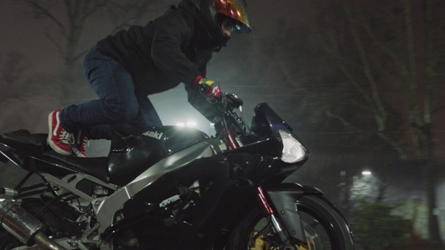 特技驾驶员骑着摩托车走向镜头，跳起来站在座位上，在雨夜做一个前倾动作。视频下载