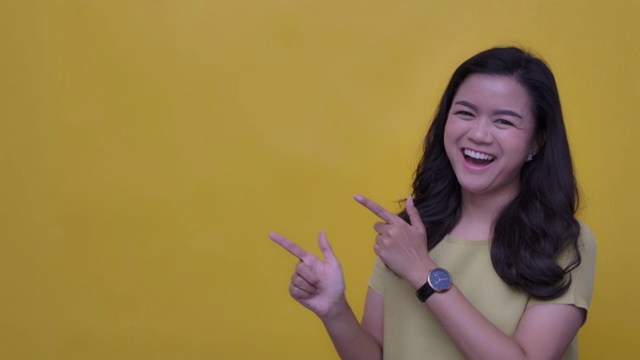 SLO MO肖像的年轻亚洲与一个幸福的微笑指向你的标志-手势在黄色的背景视频素材
