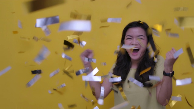 SLO MO在黄色背景慢动作拍摄的一个欢快迷人的年轻亚洲妇女庆祝派对在金色的纸屑工作室视频下载