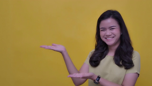SLO MO肖像的年轻亚洲与一个幸福的微笑指向你的标志-打开指向手势在黄色的背景视频素材