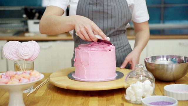 女糕点师的双手，用不锈钢刮刀和旋转蛋糕架，用粉色奶油糖霜装饰手工蛋糕，站在餐桌旁，跟踪左图视频素材