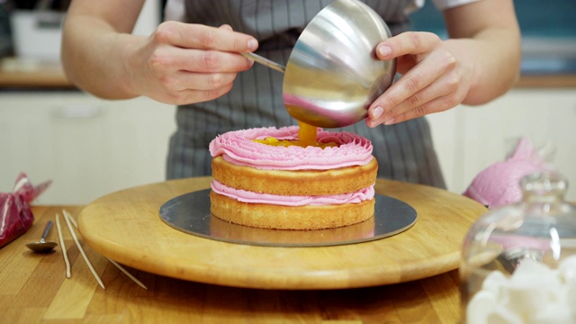 跟踪左近距离拍摄的女糕点师正在用旋转蛋糕架制作美味的海绵层蛋糕，挤出袋装粉色奶油和杏子果冻视频下载