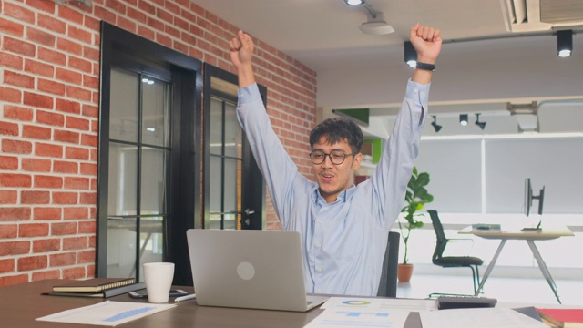 快乐的亚洲办公室的人在笔记本电脑上阅读好消息，微笑和手臂在他的办公桌在办公室，取得成功和庆祝的情绪视频素材