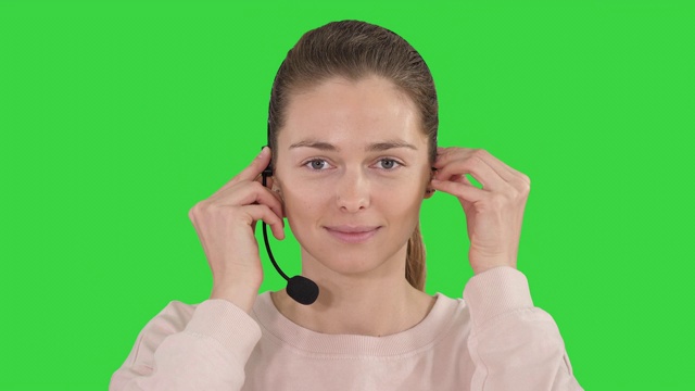 漂亮的女性支持中心操作员戴着耳机对着摄像头微笑，绿色屏幕，色度键视频素材
