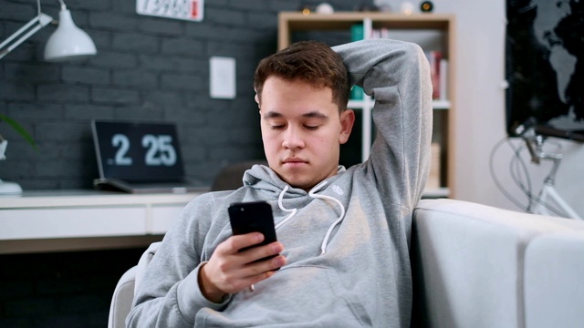 一个穿着连帽衫的男孩舒服地躺在沙发上，用他的智能手机发短信视频素材