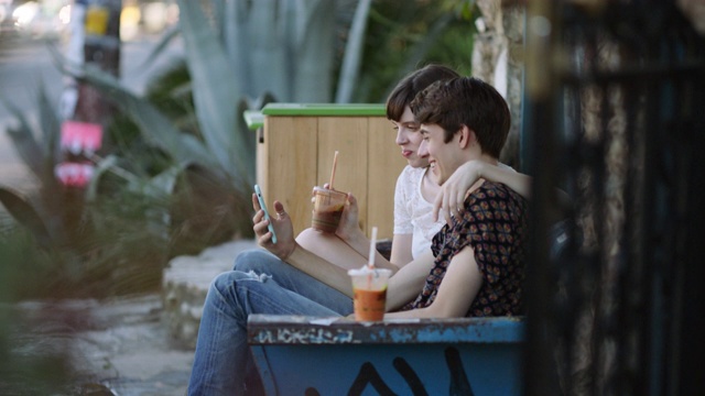 一对千禧一代潮人情侣在一家城市咖啡馆外的长椅上大笑，一边喝着冰沙，一边用iPhone视频下载
