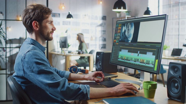胡子和牛仔裤衬衫的男性视频编辑在他的大屏幕个人电脑上工作。他在一个很酷的阁楼办公室工作。其他有创意的女性同事走在后面。视频素材