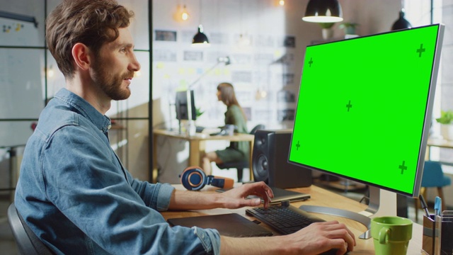 留着胡子、穿着牛仔裤衬衫的男性视频编辑在他的个人电脑上工作，电脑上有绿色大屏幕模拟显示。他在一个很酷的阁楼办公室工作。其他有创意的女性同事走在后面。视频素材