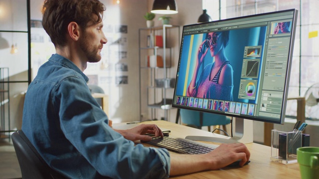 穿着牛仔裤衬衫的男性数码编辑在他的大屏幕个人电脑上使用照片编辑软件。他在很酷的办公室阁楼工作。其他有创意的女性同事走在后面。视频素材