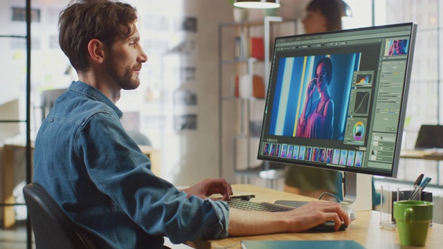 穿着牛仔裤衬衫的男性数码编辑在他的大屏幕个人电脑上使用照片编辑软件。他在很酷的办公室阁楼工作。其他有创意的女性同事走在后面。视频素材