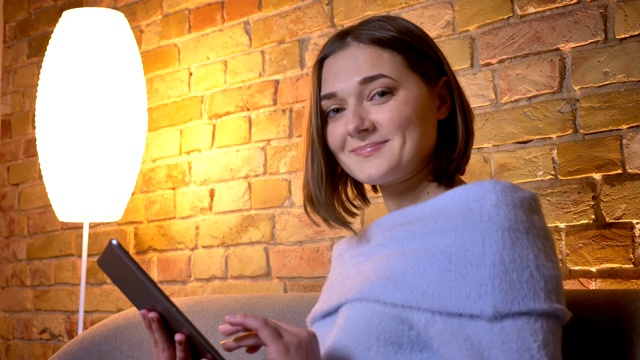 年轻美丽的白人女性使用平板电脑的特写镜头，然后看着相机，微笑，而坐在舒适的家里的沙发室内视频素材
