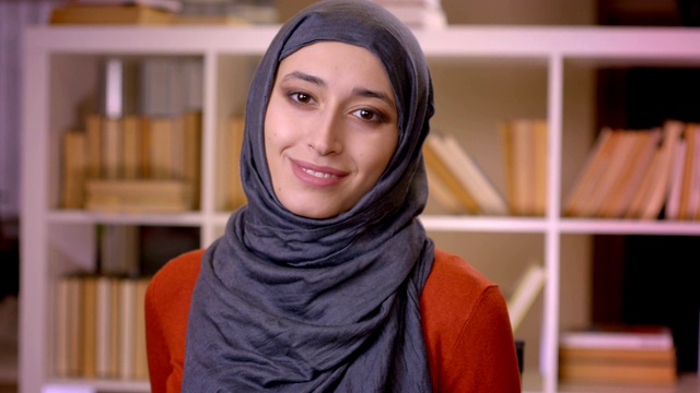 年轻貌美的穆斯林女学生戴着头巾在图书馆里看书，然后看着相机的特写镜头视频下载