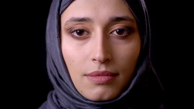特写镜头年轻迷人的穆斯林女性的脸戴着头巾，她的眼睛闭上，然后看着相机与背景孤立在黑色视频下载