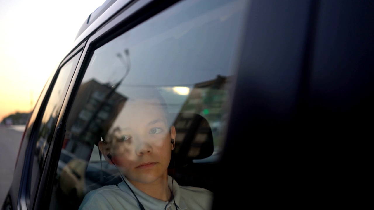 十几岁的男孩戴着耳机在汽车后座听音乐，然后看向别处。视频下载