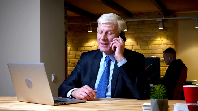 特写拍摄的老白人商人有一个电话愉快地微笑，而使用笔记本电脑在室内的办公室工作地点视频素材