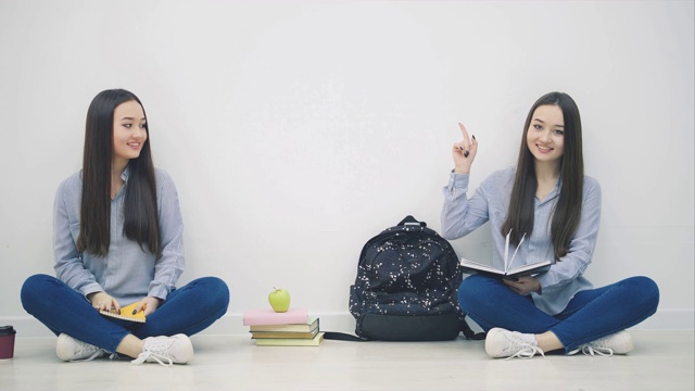 两个可爱的亚洲女孩坐在地板上，摆出莲花的姿势，互相挥手打招呼，竖起手指，竖起大拇指，微笑着。视频下载