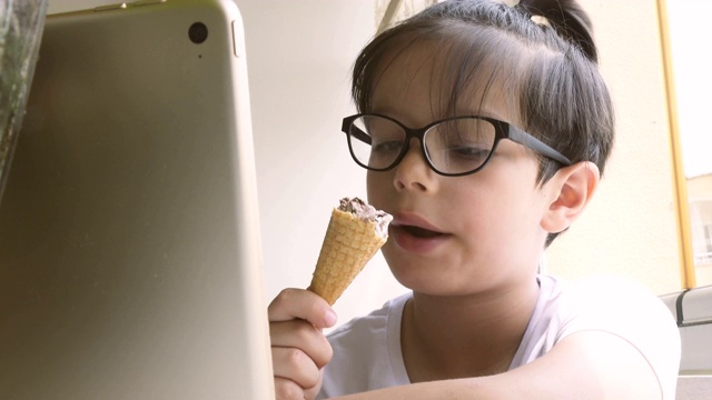 可爱的男孩舔着蛋筒里的冰淇淋，用着平板电脑视频素材