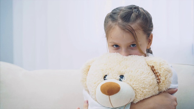 梳着辫子的快乐小女孩坐在沙发上，紧紧地抱着一只白色的大泰迪熊。视频素材