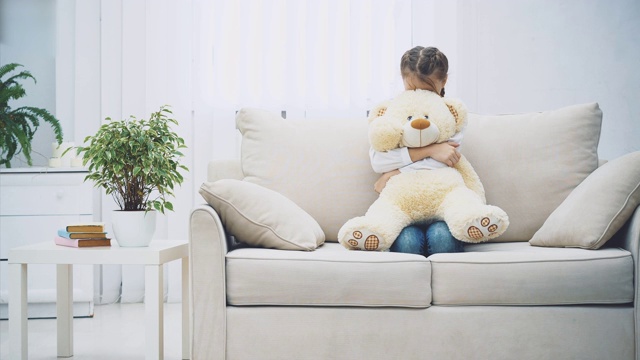 快乐的小女孩蜷着腿坐在沙发上，抱着一只大泰迪熊。视频下载