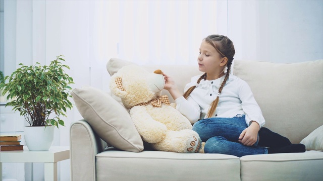可爱的小女孩坐在沙发上，和她的泰迪熊聊天，在它耳边低语着一些秘密。视频素材