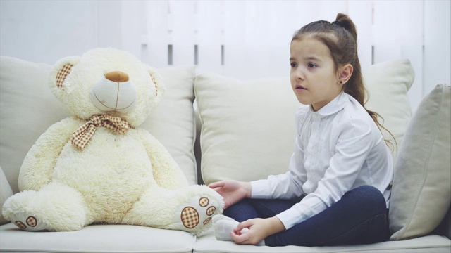 可爱的小女孩坐在沙发上，和她的泰迪熊聊天，绝望地解释着，交叉着双手，看起来被冒犯了。视频下载
