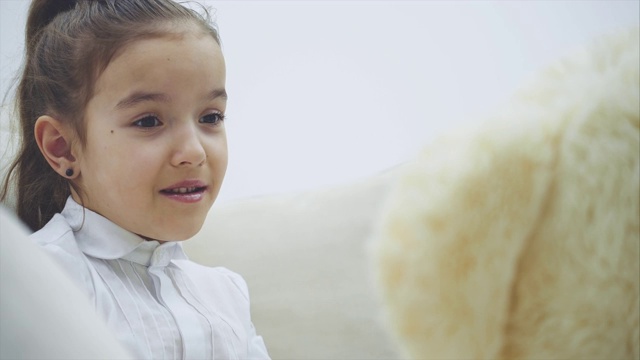 漂亮的小女孩坐在沙发上，和她的白色泰迪熊说话。视频下载