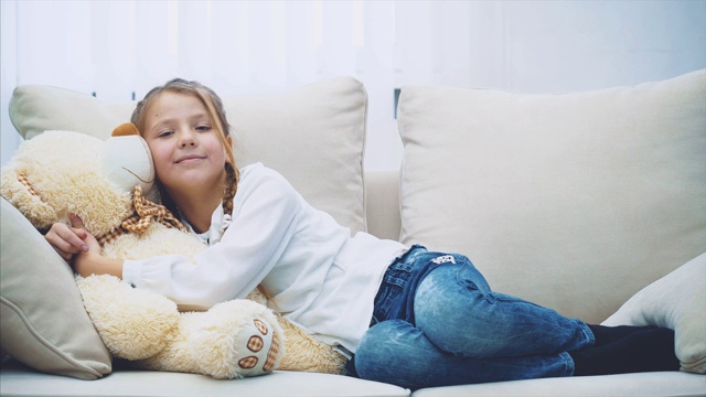 可爱的小女孩躺在沙发上，抱着她的大泰迪熊。视频下载