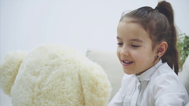可爱的小女孩坐在沙发上，和她的泰迪熊聊天。视频下载