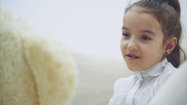 可爱的小女孩坐在沙发上，和她的泰迪熊聊天。视频素材