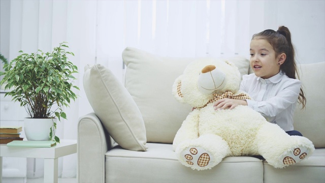 可爱的小女孩坐在沙发上，和她的泰迪熊聊天，在它耳边低语着一些秘密。视频下载