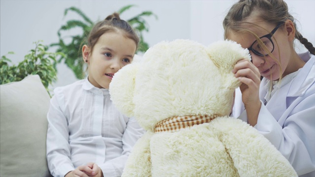 两个可爱的孩子在玩泰迪熊。熊病了。一个女孩是医生，正在检查毛绒熊。另一个女孩是熊的主人。视频下载