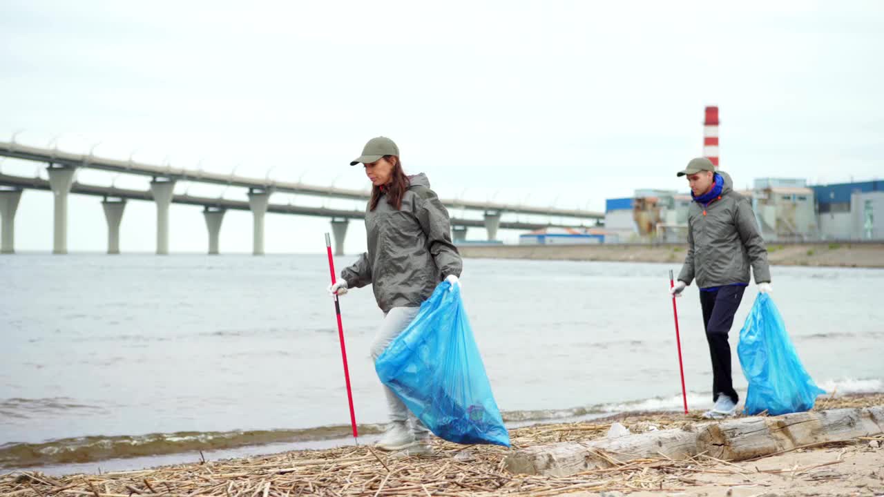 左镜头是两个环保人士，男人和女人拿着拾荒器和垃圾袋沿着海岸线走，在肮脏的海滩上捡垃圾视频下载