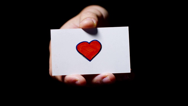 一只手拿着一张卡片，上面画着一颗孤立在黑色背景上的心。视频素材