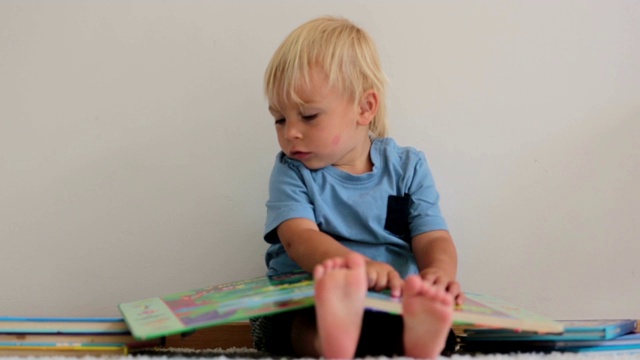 可爱的蹒跚学步的男孩，在家里看书，坐在地上，他周围有很多书视频下载