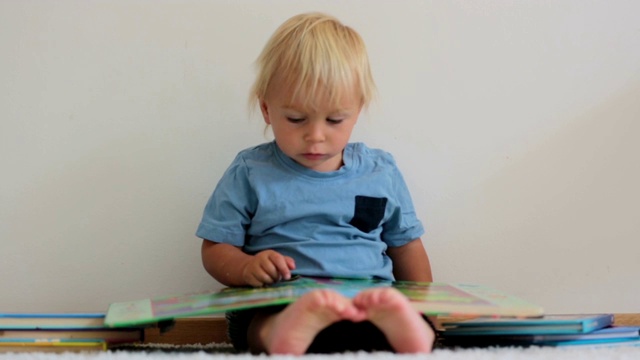 可爱的蹒跚学步的男孩，在家里看书，坐在地上，他周围有很多书视频素材