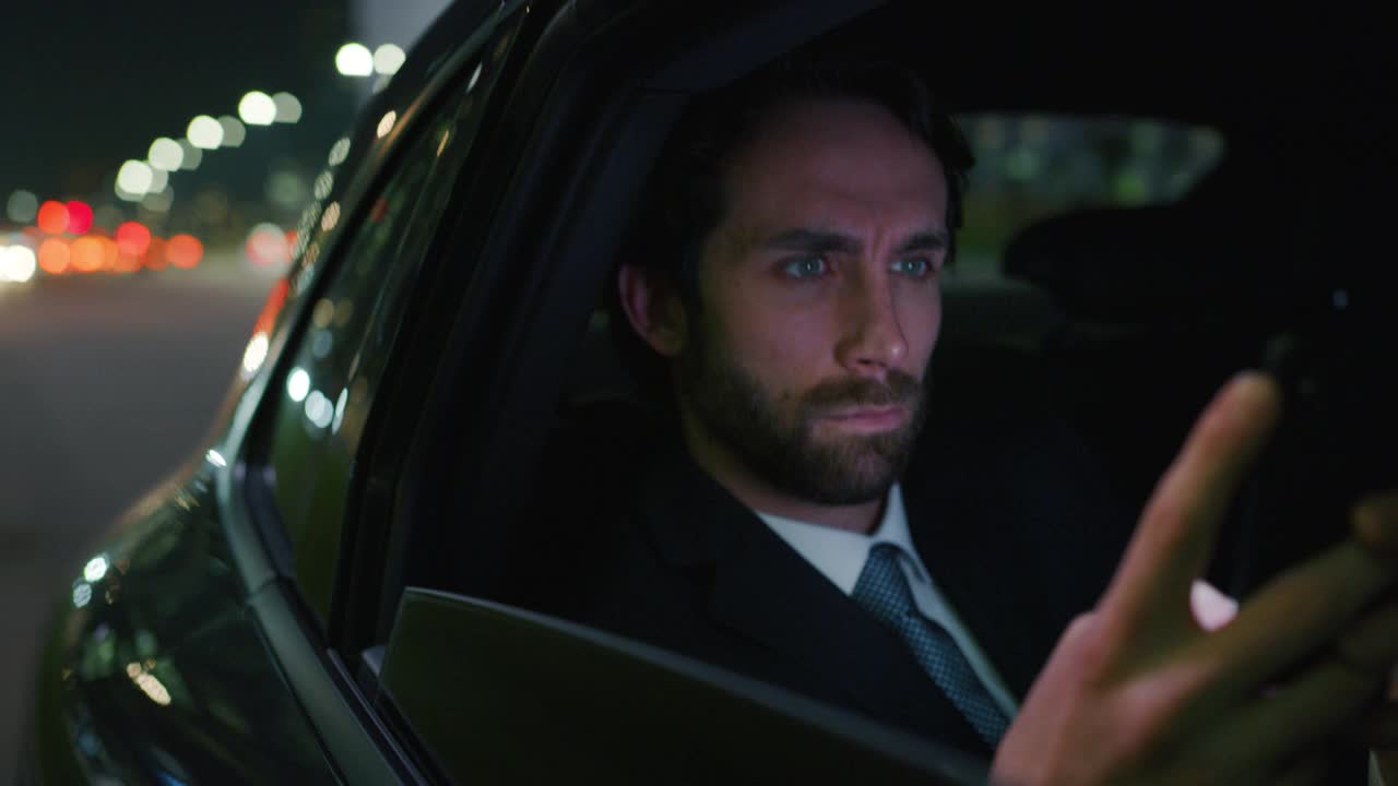 一个英俊的商人用他的笔记本电脑在一辆现代汽车和司机在城市中心的夜晚旅行的慢镜头。视频下载