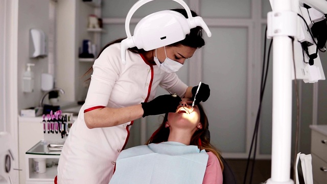 牙医办公室，一名年轻女牙医穿着白袍和口罩检查和工作的病人的牙齿，指导光设备的嘴。牙科保健视频素材
