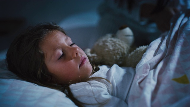 年轻女子的母亲给她的小女儿一个晚安吻，而她正睡在床上的泰迪熊。视频下载