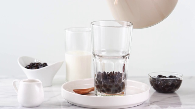将奶茶倒入玻璃杯中，配以台湾流行的木薯珍珠泡，放在明亮的大理石桌上和白色托盘上，这是自制的概念。4 k视频拍摄。视频素材