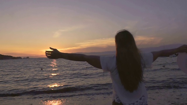 一个女人在夕阳下张开双臂站在海滩上视频素材