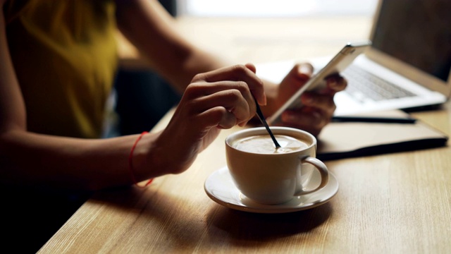 女人的手在杯子里搅拌咖啡和使用智能手机在咖啡馆放松的特写视频素材