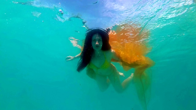 美丽的年轻女孩在黄色泳衣与飘动的织物和长头发摆姿势水下。她游着，胳膊伸着，看着镜头。4 k。视频下载