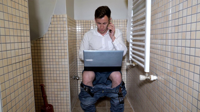 一个年轻的工作狂在厕所里用电脑工作，用智能手机聊天。视频下载