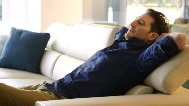 一个男人躺在家里的沙发上，抱着一个枕头，微笑着看帆布电影。视频下载