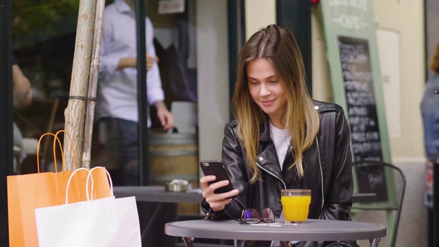 购物后，年轻的女子正在酒吧吃早餐，而短信她的朋友与手机和笑得开心。视频素材