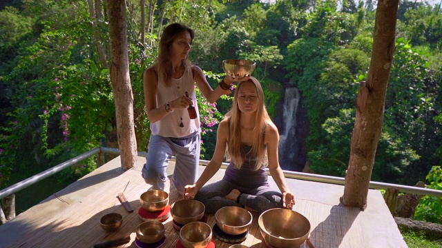 一名精通亚洲圣药的女子为一位年轻女性客户表演藏式碗治疗仪式的超慢镜头。用西藏唱碗冥想。他们在一个凉亭里冥想，背景是美丽的瀑布视频素材