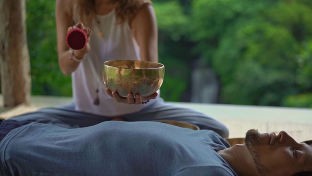 一名精通亚洲圣药的女子为一名年轻男子执行藏碗治疗仪式的超慢镜头。用西藏唱碗冥想。他们在一个凉亭里冥想，背景是美丽的瀑布视频素材