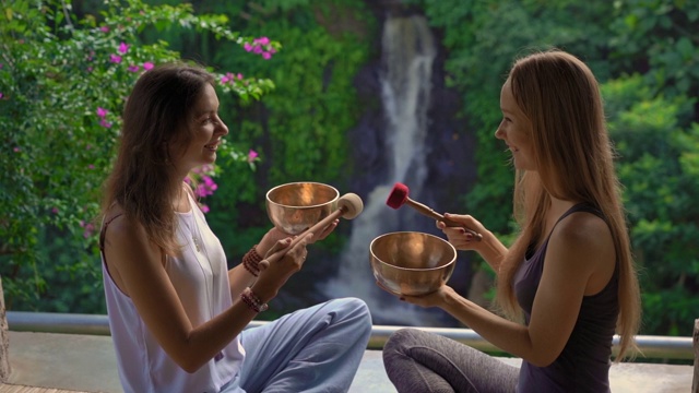 一名精通亚洲圣药的女子为一位年轻女性客户表演藏式碗治疗仪式的超慢镜头。用西藏唱碗冥想。他们在一个凉亭里冥想，背景是美丽的瀑布视频素材